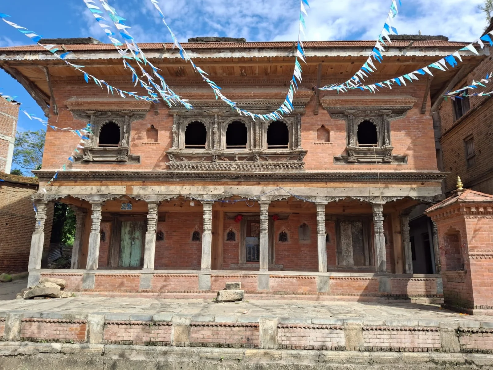 The finished Etay community building: a two-storey red-brick building with carved wooden windows, a row of carved wooden pillars along the ground floor and a raised stone platform in front, under strings of blue and white bunting