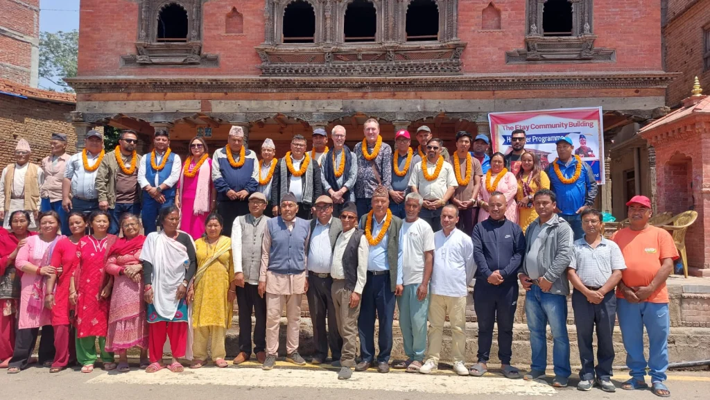 Villagers, local officials and ARNF supporters, many wearing marigold garlands, standing together in front of the new building under a banner reading The Etay Community Building Handover Programme