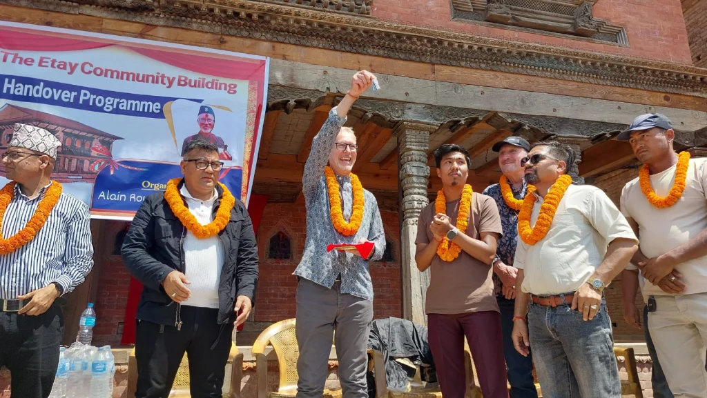 A man in a marigold garland smiles and holds up a set of keys at the handover, with local officials in garlands beside him and the handover banner behind