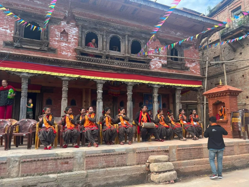 Young musicians in matching traditional dress play flutes and a drum on the platform in front of the building, while a woman watches from a carved upstairs window