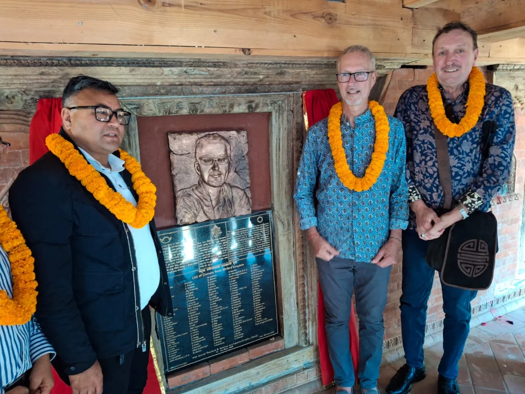 Three men in marigold garlands stand beside a newly unveiled plaque: a copper relief portrait above a black stone tablet listing donors from the United Kingdom