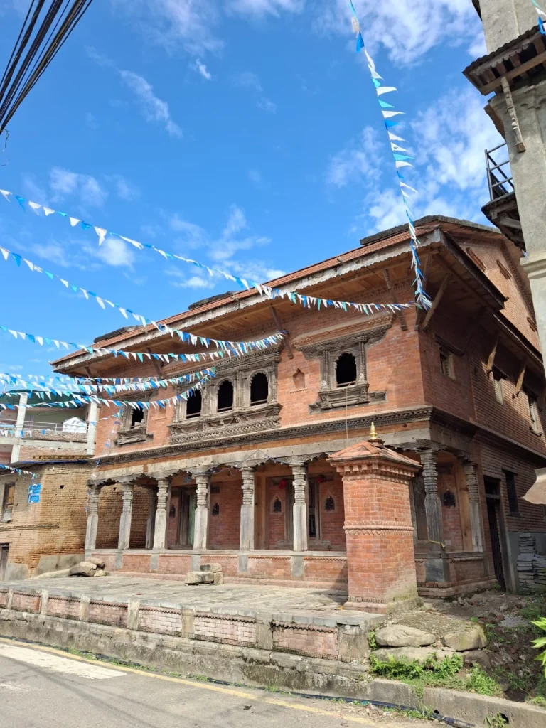 The finished building seen from the street at an angle, with its carved pillars, a small brick shrine at the corner and bunting strung overhead